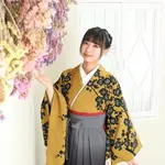 A woman in a yellow kimono and black hakama with her hand on a purple flower.