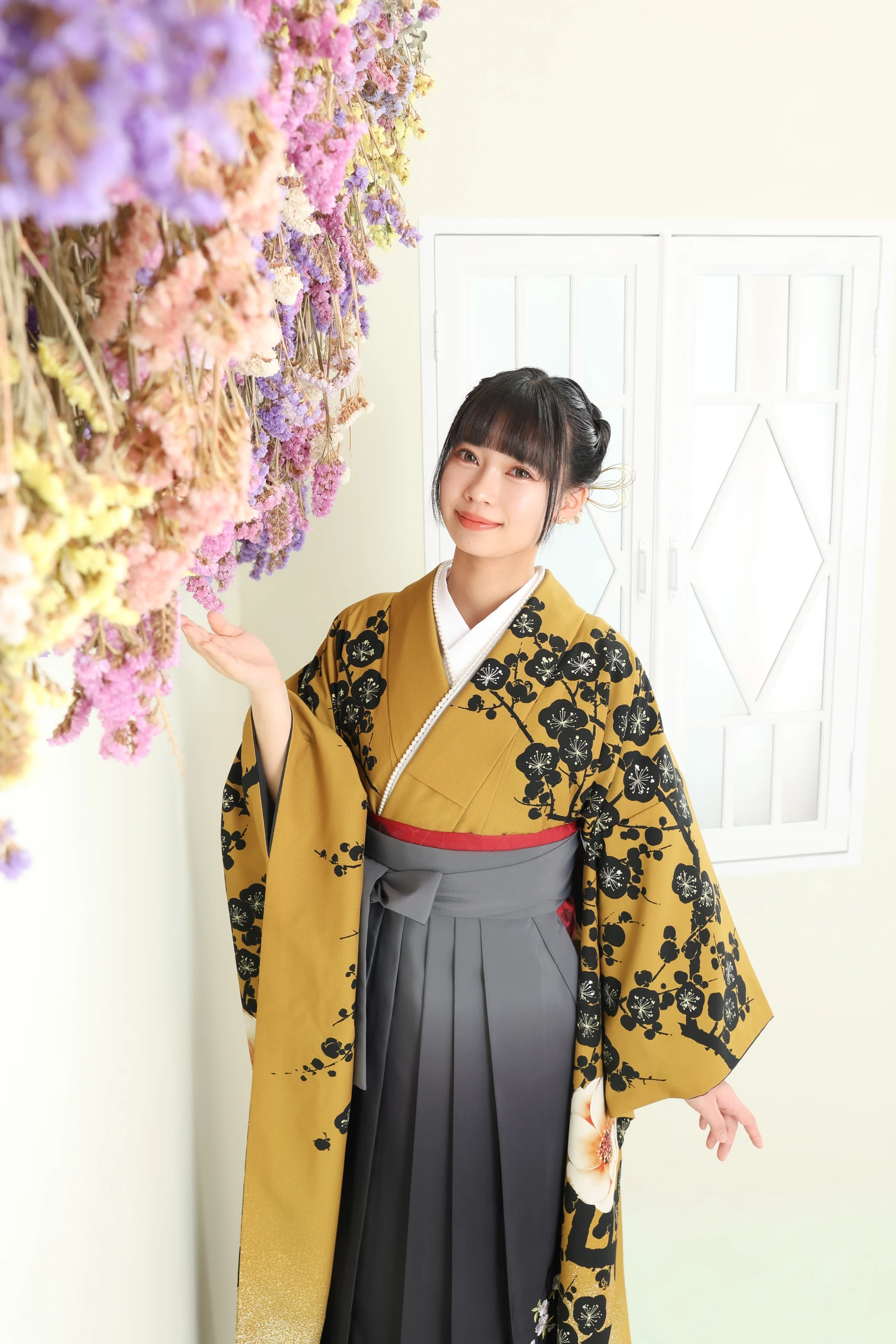 A woman in a yellow kimono and black hakama with her hand on a purple flower.
