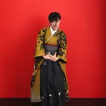 A woman in a yellow kimono and black hakama holding a graduation certificate against a red background