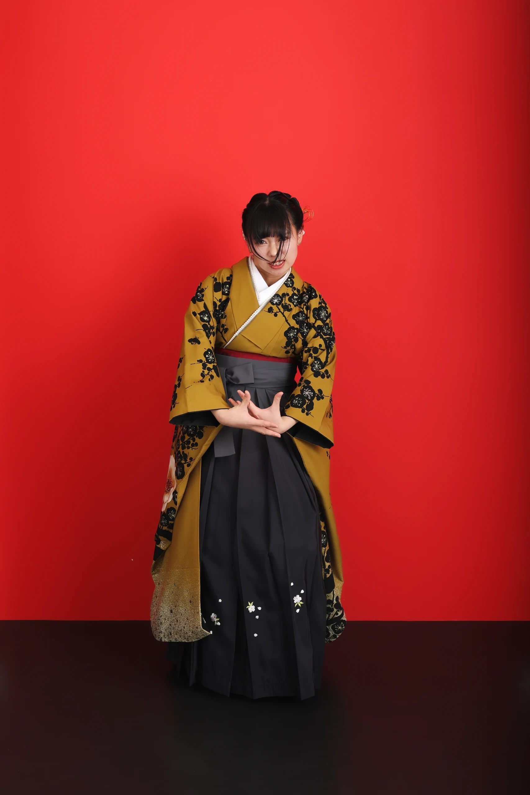A woman in a yellow kimono and black hakama holding a graduation certificate against a red background