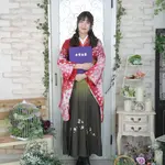 A woman in a red kimono and black hakama holding a diploma in a bright indoor set.