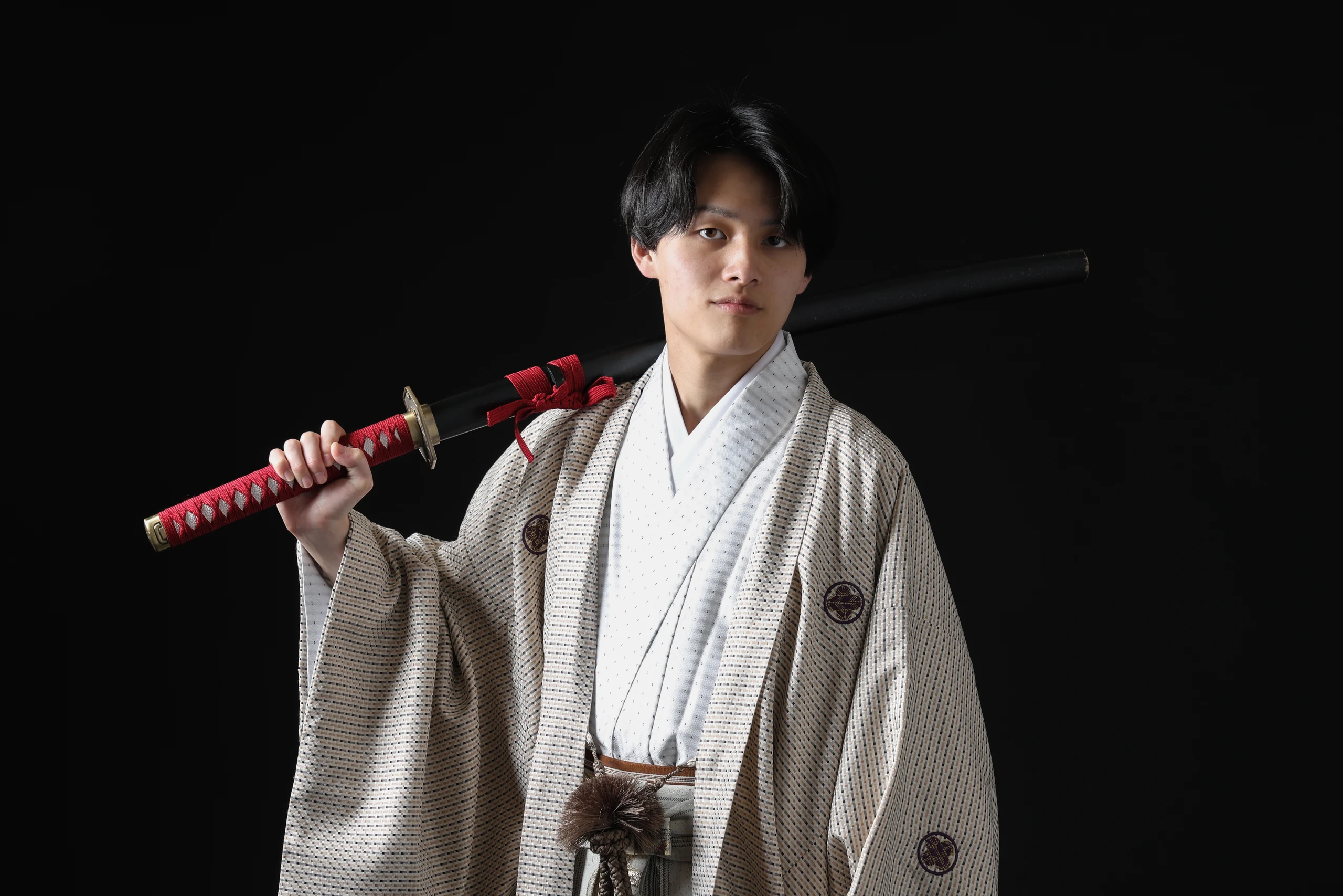 黒背景で赤い柄の刀を肩に担ぐ淡色紋付袴の成人男性
