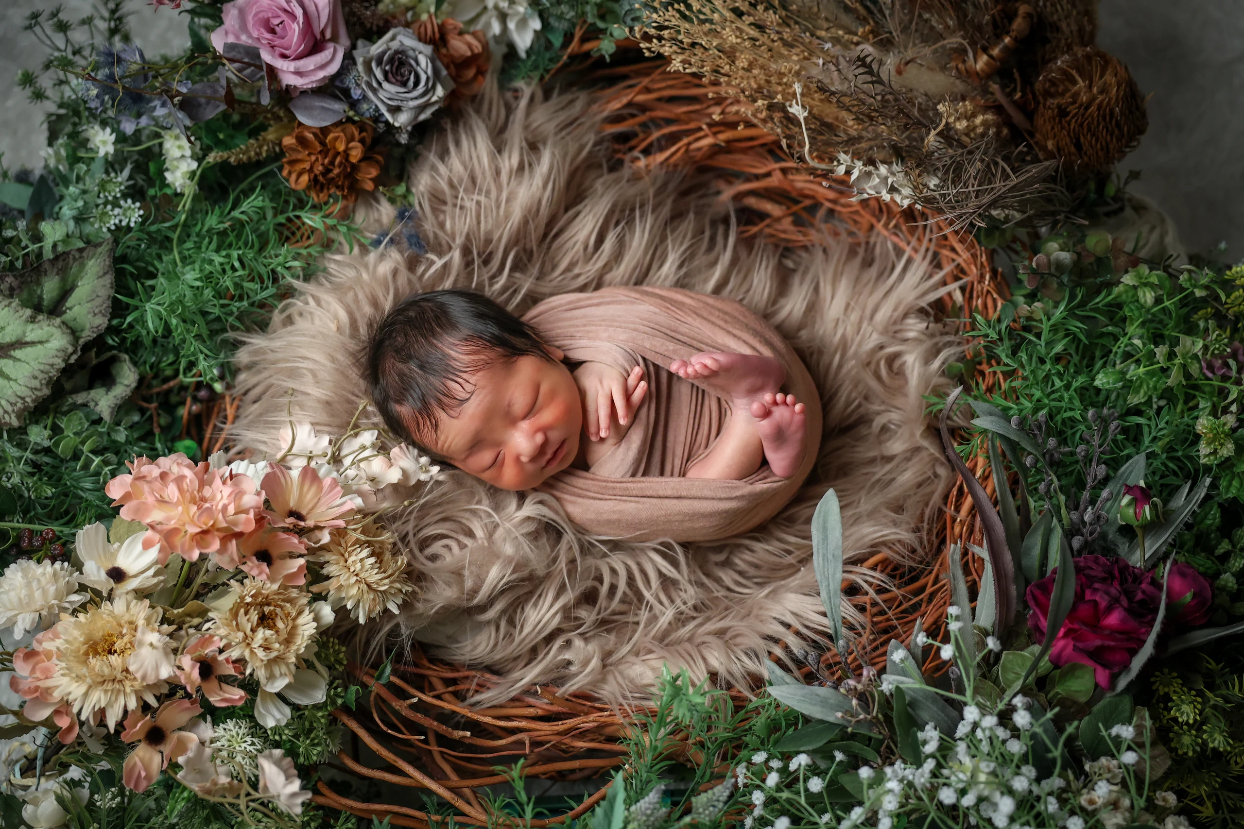 茶色のおくるみに包まれ花とグリーンの中で眠る赤ちゃん