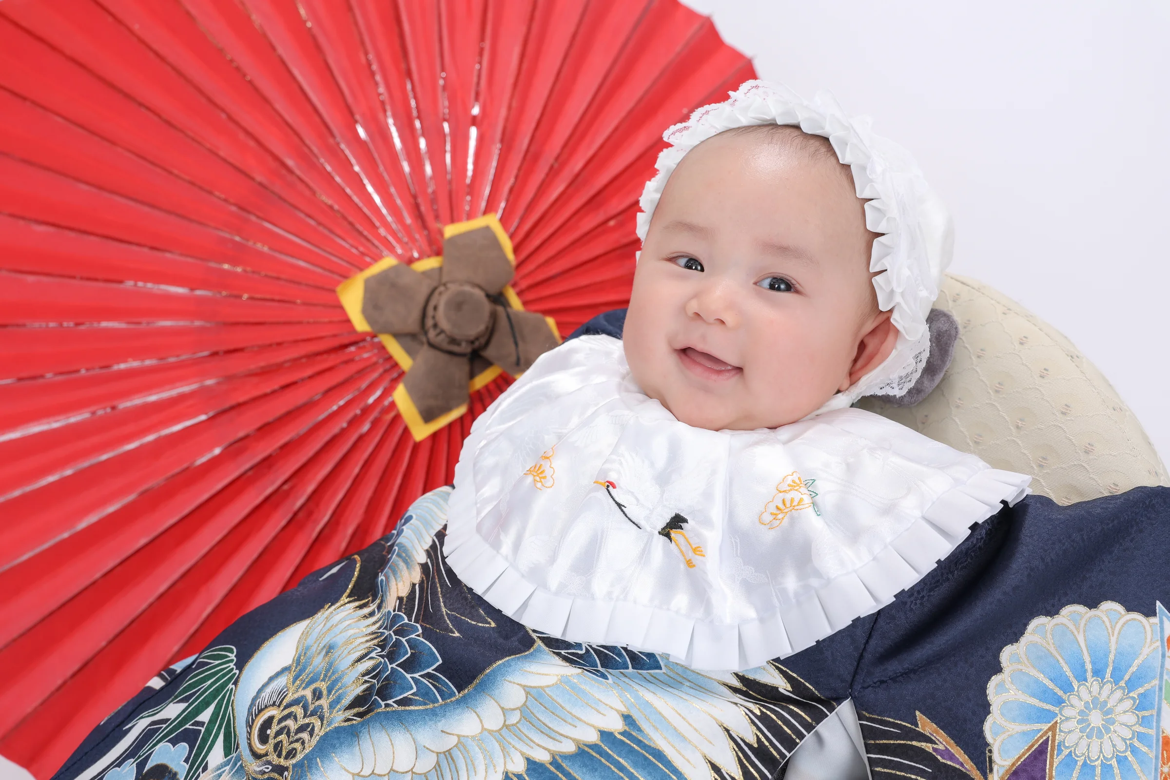 紺色の祝い着をまとい赤い和傘を背景に笑う赤ちゃん