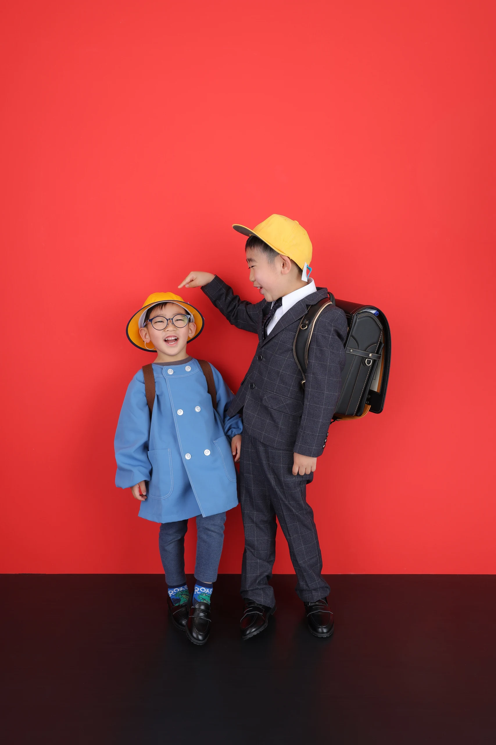 赤い背景で黄色い帽子とランドセルを見せるきょうだい