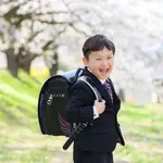 A boy smiling while carrying a black backpack under full-bloomed cherry blossoms