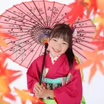 A seven-year-old girl smiling with autumn leaves in the foreground bokeh and a pink Japanese umbrella in the background.