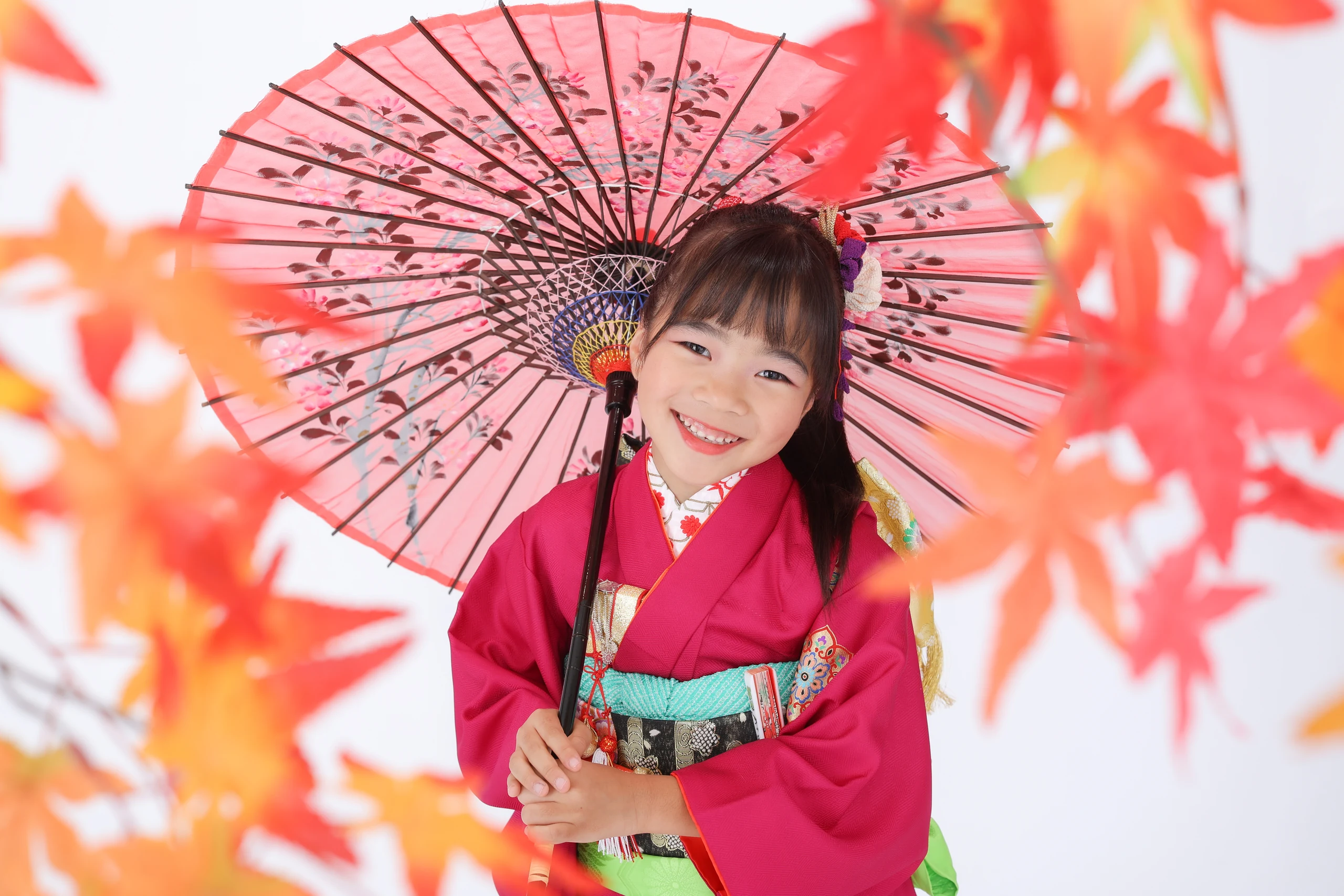 紅葉の前ボケとピンクの和傘を背景に笑う七歳の女の子