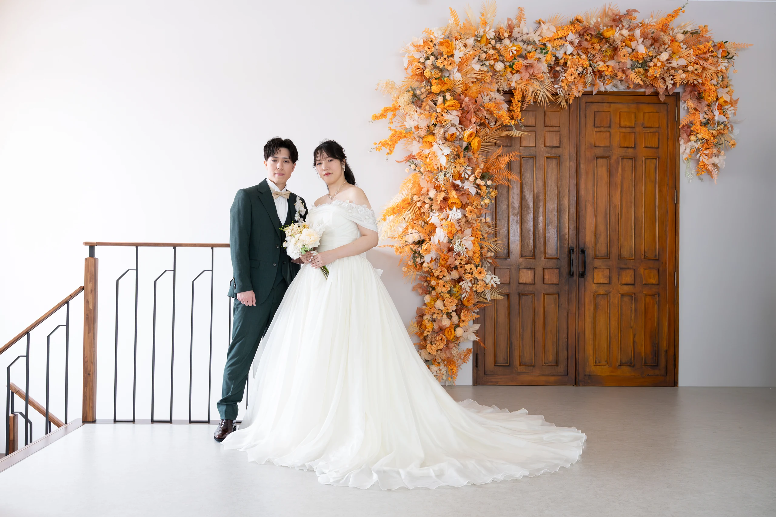 オレンジ色の花で飾られた木製ドアの前に立つウェディングドレスと深緑のタキシード姿の新郎新婦