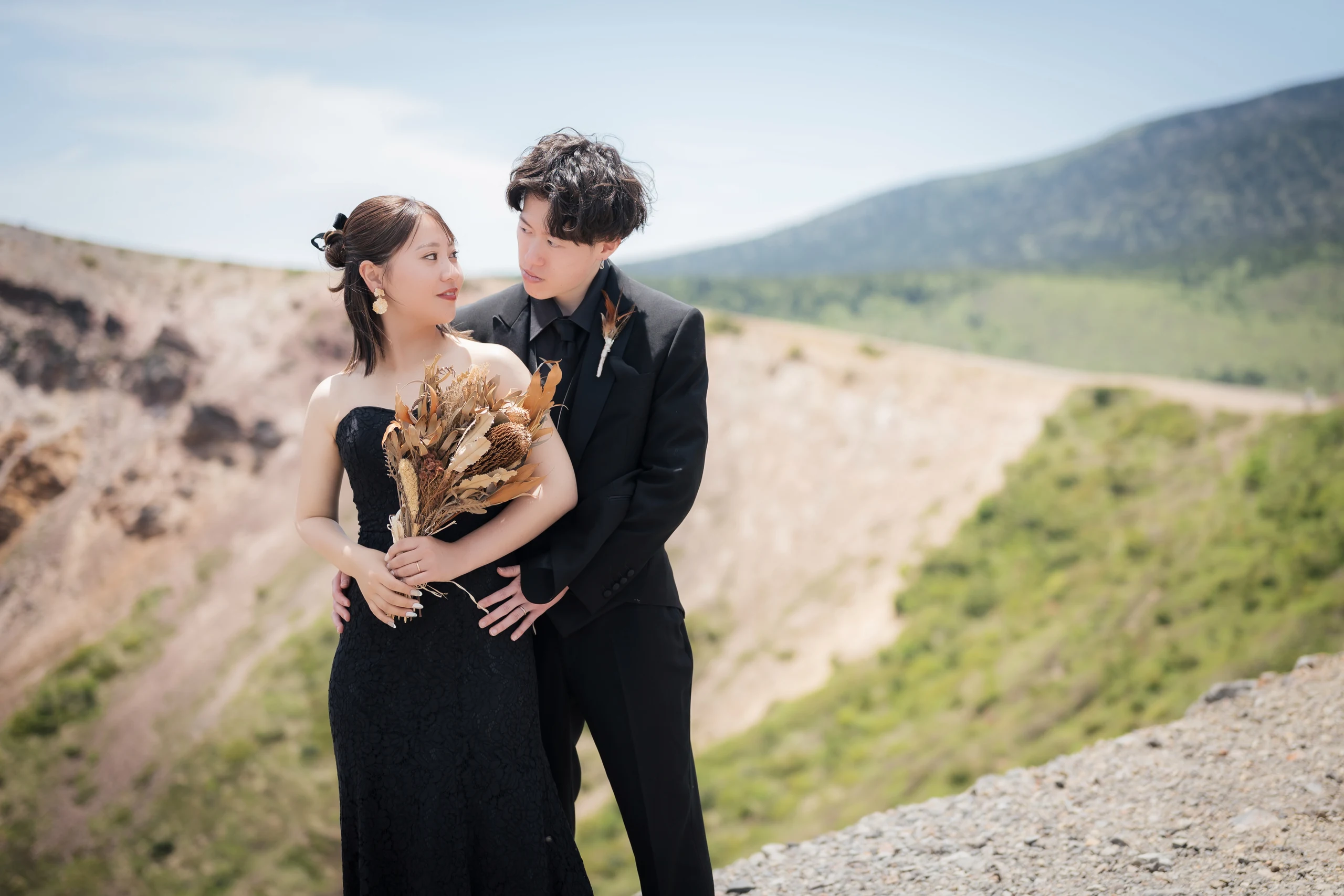 山の景色を背景に黒いドレスとタキシード姿で寄り添う新郎新婦