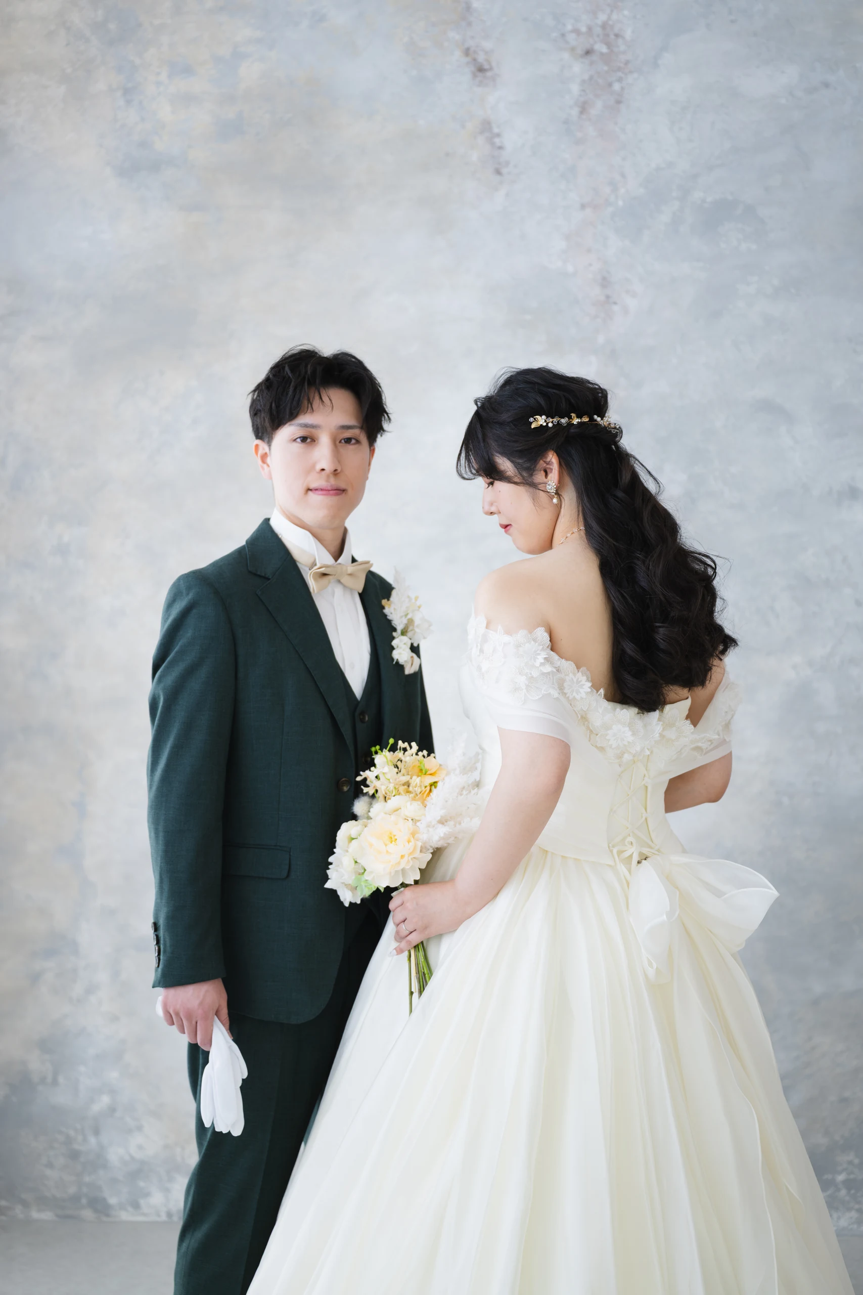 グレーの背景で白いウェディングドレスと深緑のタキシード姿の新郎新婦