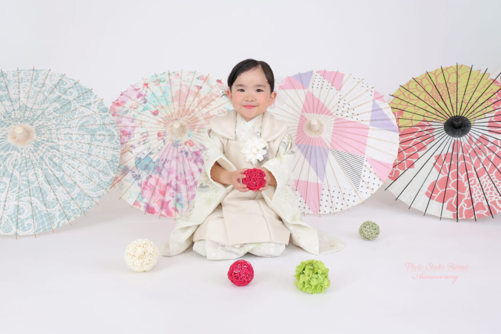 和傘と毬を背景に座る、淡い色の着物姿のお子さま
